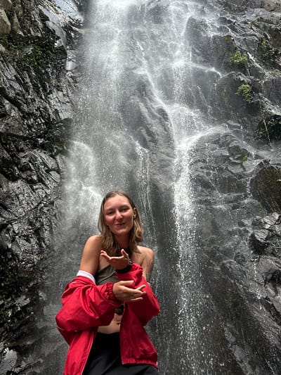 The image captures a young adult female posing in front of a waterfall in Tu Le, Vietnam. The subject, positioned centrally, has light-colored skin, long light brown hair, and is looking directly at the viewer with a smile. She is wearing a dark olive green strapless top, a bright red long-sleeved jacket unzipped and worn off her shoulders, exposing her midriff, and dark-colored bottoms. Her hands are held in front of her chest, with fingers loosely curled and palms slightly open, as if in an expressive gesture.
Behind the woman is a large, cascading waterfall flowing over a dark, rugged rock face. The rock is dark grey to black, appears wet and textured, and is interspersed with patches of green foliage, including ferns and small plants, particularly visible on the left side of the frame and in smaller clusters throughout the rock face. The waterfall itself consists of numerous streams and sprays of white water, creating a dynamic and misty background. The upper portion of the waterfall and cliff extends out of the frame. The overall scene depicts a natural, outdoor environment.