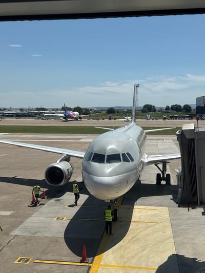 An Airbus A320 aircraft, painted white with Arabic script on its fuselage, is parked at a gate at Nikola Tesla Airport in Surcin, Serbia. The aircraft is connected to a jet bridge, and several ground crew members wearing high-visibility vests are visible on the tarmac, attending to the plane. One crew member is placing a traffic cone, while others appear to be performing routine checks. In the background, another aircraft, a Wizz Air Airbus A321, is visible on a taxiway. The scene is set under a clear blue sky with scattered clouds, indicating fair weather conditions. The airport environment is characterized by concrete tarmac, painted markings, and various airport infrastructure like terminals and service vehicles.