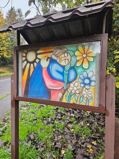 A medium shot captures a framed painting under a small shelter. The painting depicts two figures, one with green hair, embracing against a backdrop of colorful flowers and a stylized sun. The shelter has a brown metal frame and corrugated roof. The immediate environment around the shelter includes grass, fallen leaves, and foliage. The presence of wet leaves and sheen on the frame suggests it has recently rained. The location, Osorno, Chile, is assumed based on context.