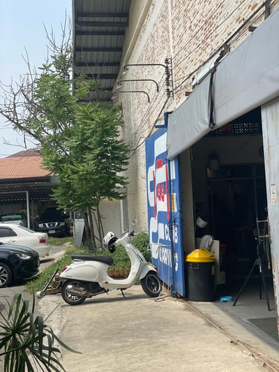 The image shows a street scene in Ban Rong Ruea Kham, Thailand. A white scooter is parked on the sidewalk, next to a black car and a white car. A tree partially obscures the view of a building behind the vehicles. A large blue and white container, possibly repurposed, is positioned near the street, its side marked with writing and a logo. An awning extends from the building. There is a trash can and a helmet visible inside of the repurposed container. Power lines are strung across the frame.