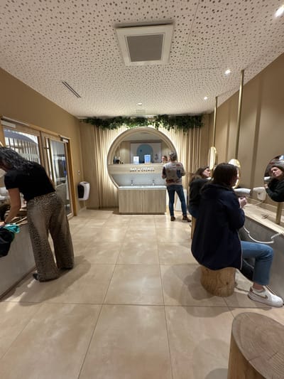 This image captures an interior view of a beauty or hair treatment facility located in Marcq-en-Barœul, France. The room features light beige walls, a light-colored tiled floor, and a white acoustical ceiling with a speckled pattern, recessed lighting, and rectangular air vents.
In the center of the background, a prominent circular wooden frame surrounds a large mirror and a "BAR À CRÈMES" sign. Below this, two stainless steel sinks with faucets are set into a light wood cabinet. Above the mirror, artificial green foliage decorates the wall. A woman in a light-colored patterned shirt and blue jeans stands facing this central counter, her reflection visible in the mirror alongside a light blue and white abstract artwork.
To the right, a long, light-colored counter extends, featuring several oval mirrors separated by vertical brass-toned rods. Multiple white hair dryers are mounted along this counter. A woman in a dark blue jacket and blue jeans is seated on a cylindrical wooden stool, facing away from the camera, holding a white hair dryer. Her reflection is visible in one of the oval mirrors. Another woman in a dark top is partially visible behind her, looking towards the central counter. Additional cylindrical wooden stools are present to the right.
On the left side of the foreground, a woman with dark, curly hair, wearing a black top and leopard print wide-leg pants, is bent over a light-colored counter, engaged with a green fabric item. Further back on the left wall, a white wall-mounted dispenser is visible, along with two light wood doors.