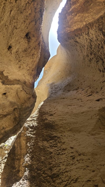 The image depicts a view looking upwards within a natural rock formation in the city of Ziarat, Pakistan. The scene shows a tight space between large sandstone-colored rock formations, possibly inside a cave or narrow canyon. Sunlight is visible through gaps in the rock ceiling, revealing a blue sky. The surfaces of the rocks are textured, with some showing evidence of weathering and erosion. There are no people or identifiable objects present. The overall impression is of a natural, geological feature.