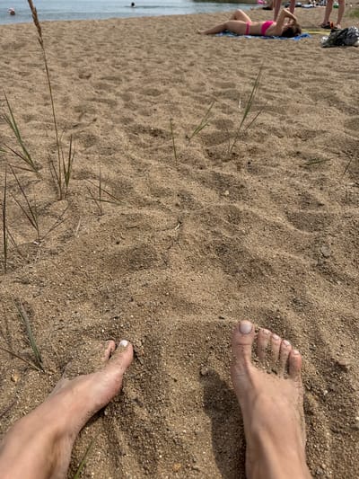 The image shows a close-up of a person's bare feet in sand on a beach. In the background, several other people are visible, some in the water and some lying on the sand. The water appears to be a lake or calm sea, and the beach is sandy with some sparse vegetation. The scene suggests a casual, recreational day at the beach. The city of Ayyrtau, Kazakhstan, is located near a large lake called Lake Ayyrtau, which is a likely location for this scene.