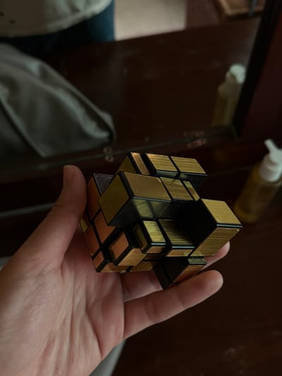 A distorted golden Rubik's Cube sits in the palm of a person's hand. The cube has an irregular shape, with the metallic gold and black pieces protruding at varying lengths. Background elements include a brown wooden surface with a bottle and a blurry gray cloth. The focus is sharp on the cube and the hand, contrasting against the blurred background.