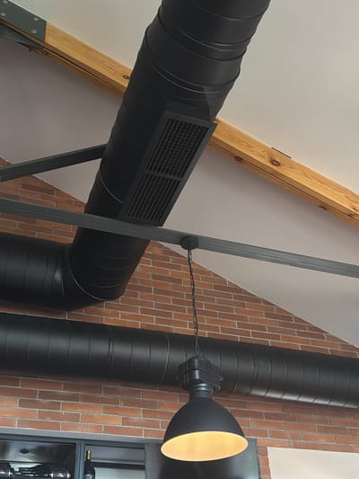In Byurakan, Armenia, this image captures an interior view of a building's exposed ceiling. The ceiling features prominent black industrial-style ventilation ducts that run horizontally across the frame. These ducts are supported by dark gray metal beams, which are also attached to a light-colored pitched roof. A wooden beam, likely structural, runs diagonally across the upper portion of the image.
Visible in the lower part of the image are sections of exposed red brick wall. Hanging from one of the metal support beams is a black industrial pendant light with a wide, inverted bell-shaped shade that is illuminated from within. The overall aesthetic suggests a modern, industrial design, possibly in a restaurant, cafe, or open-plan office space. There are no people or animals visible in the image.