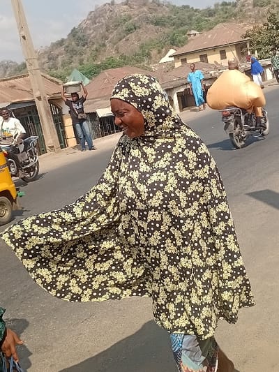 An elderly woman, clad in a black floral-patterned hijab and dress, occupies the foreground of a street scene. In the background, a sandy-colored mountain rises behind a row of modest buildings. Various people are visible: some stand by the buildings, while others ride motorcycles, one carrying a large, wrapped object. The scene is bathed in bright daylight.