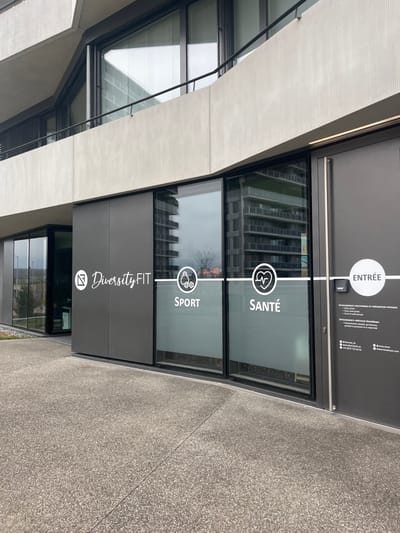 The image displays the modern, glass-fronted entrance of a business named "DiversityFIT" in Meyrin, Switzerland. The shot is taken from an outdoor, ground-level perspective, looking towards the building's facade.

The entrance features a dark grey door and large glass panels, reflecting the surrounding urban environment, including adjacent buildings with numerous windows and a bare tree against an overcast sky. The ground in front of the building is a speckled grey aggregate pavement.

White vinyl graphics are prominently displayed on the door and windows. A vertical white line runs down the center, intersected by a horizontal line. At the top, a circle contains the word "ENTRÉE" (French for "Entrance"). To the right, smaller text in French lists services and contact information: "ACCOMPAGNEMENT ET PRÉPARATION PHYSIQUE" (Accompaniment and Physical Preparation), individual, semi-private, and group classes, physical preparation, individualized programming, assessments & advice, and natural food supplements. Contact details include "@diversityfit", "www.diversityfit.ch", and a Swiss phone number.

Below the "ENTRÉE" sign, on the glass panels, are two icons within white circles, labeled in French: a heart/pulse icon with "SANTÉ" (Health) and a bicep with a dumbbell icon with "SPORT". Further down, on a solid dark panel, the business name "DiversityFIT" is written in a script font, accompanied by a stylized logo (possibly a 'D' or a delta symbol) in a white circle.

The overall scene suggests a contemporary health and fitness center, characterized by clean lines and functional design, typical of a modern commercial establishment during daytime. No people are visible, and no specific activity is taking place at the moment of the photograph.