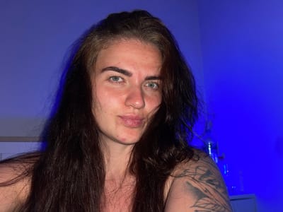 A medium shot photo shows a young woman with long, dark brown hair and light green eyes. She is looking directly at the camera with a neutral expression. Her skin is fair and shows some redness and blemishes around her cheeks and chin. She has prominent eyebrows and her lips are slightly parted. She is wearing a dark, possibly strappy top, and a tattoo is visible on her right shoulder. The background is a solid, vibrant blue, with a hint of a white object or wall on the left side and some indistinct objects on the right. There is no indication of the location being Мосметаллоконструкция Онт, Russia, or any urban environment.