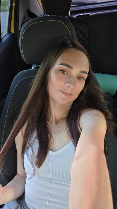 A young woman with long brown hair is taking a selfie from the driver's seat of a car. She is wearing a white ribbed tank top. The interior of the car is visible, including a black seat with yellow stitching, and a yellow exterior panel. The scene appears to be outdoors, possibly on a street in Bulgakovo, Russia, given the context.