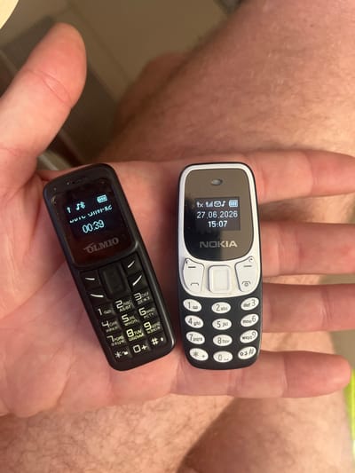 The image displays a close-up of a person's hand holding two small mobile phones. The top phone is a white and black Nokia feature phone, clearly displaying the date "27.06.2026" and the time "15:07" on its screen. The bottom phone is a black feature phone with the brand name "OLMIO" visible on its screen, showing "00:39" as the time. The background of the image is out of focus, but appears to be a person's skin, possibly an arm, with visible hair. The lighting suggests it is daytime. The image is a still photograph, with no specific activity or event other than the phones being held. The location context provided is "Balaklavsky District, Ukraine," but there are no visual cues in the image to confirm this location. The overall impression is a simple capture of two retro-style mobile phones in a person's hand.