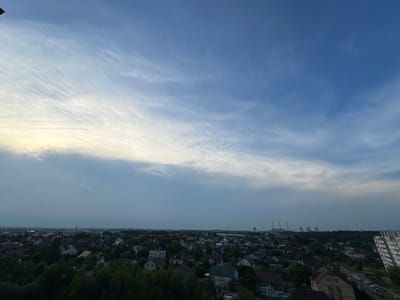 This is an aerial view of a suburban area in Шчомысліцкі сельскі Савет, Belarus, likely taken during the late afternoon or early evening, given the soft lighting and the sky's appearance. The scene is dominated by a vast expanse of sky, featuring a gradient of blue with wispy, white clouds streaking across the upper portion, transitioning to a more overcast and muted gray near the horizon.
Below the sky lies a sprawling residential area characterized by numerous houses with varied rooflines and colors, interspersed with dense green trees and vegetation. The buildings appear to be mostly detached homes, suggesting a suburban or semi-rural setting. In the distance, several industrial structures, including tall chimneys, are visible, indicating the presence of a power plant or similar industrial facility. A modern high-rise apartment building stands out on the right side of the frame, contrasting with the more traditional housing. The overall impression is one of a quiet, residential landscape under a dynamic sky. No people or specific activities are discernable in the image. No visible text is present.
