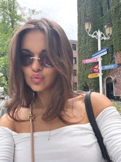 The image depicts a young woman with brown hair, wearing sunglasses, and a white mesh top with a black strap. She is making a kissing gesture. The background features greenery-covered walls, a brick building, and a signpost with directions. The signpost indicates locations in Zhangjiajie, China, including "Zhangjiajie Mr Sean Castle", "Popular Photography Sanctuary", "7th Floor Western Restaurant", "7th Floor Sky Garden", "Stay in Tianmen Mountain View Room", and "Sky Mirror on the top floor". There are also street lamps on the signpost. The overall environment appears to be an outdoor tourist area in Zhangjiajie.