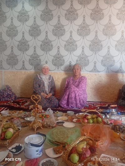 In an indoor setting, two women are seated on the floor, facing the camera. The woman on the left wears a patterned dress and a light-colored headscarf. The woman on the right is dressed in a purple patterned dress. Both are seated on a rug that covers the floor. Behind them, the wall is covered with ornate, light-colored wallpaper. In front of them, a table is laden with food and drinks, including baskets of fruit, candies, snacks, and beverages. The image captures a moment of what appears to be a meal or gathering, with the presence of the date stamp "16.07.2026 14:34" suggesting a specific time of occurrence. The location is identified as Чоң-Гара, Kyrgyzstan.