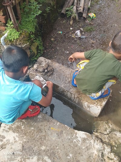 Two young boys are squatting on the edge of a concrete water channel, peering into the murky water. The boy on the left wears a light blue shirt and red sandals, while the other is in a green shirt and blue flip-flops. Trash litters the muddy ground behind them, near some stacked bamboo poles and a brick wall covered in vegetation. The scene is captured from an overhead perspective, showcasing the boys' engagement with their surroundings.
