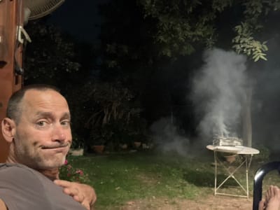 This image depicts an outdoor evening scene in a backyard located in the Brener Regional Council, Israel. In the left foreground, a middle-aged male with short dark hair and a grey t-shirt is captured in a close-up, looking directly at the camera with a slight smirk and pursed lips. To his left, a section of a reddish-brown wall or structure is visible, with a white oscillating fan mounted high. In the mid-ground, a small, square barbecue grill is placed on a white, round metal folding table. The grill is actively producing a dense, vertical plume of white-grey smoke. The background consists of a grassy lawn, several potted plants, and dark green trees and foliage silhouetted against the night sky. A bare foot is partially visible in the lower right corner, resting on a dark object, possibly part of a chair.