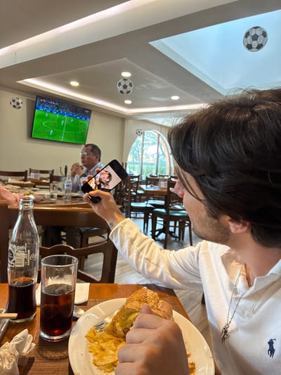 The image depicts an indoor scene, likely a restaurant or cafe, with a soccer match being broadcast on a television mounted on the ceiling. Several decorative soccer balls are also suspended from the ceiling. In the foreground, a man with dark hair is holding up a smartphone, taking a picture or recording a video. The phone screen displays a photograph of a person. In front of the man is a table with food and drinks, including a sandwich, potato chips, and two glasses of dark soda. A partially visible bottle of Coca-Cola is also on the table. In the background, another person, an older man wearing glasses, is seated at a table, looking towards the television. The environment appears to be moderately lit, with wooden tables and chairs visible, and large arched windows in the background suggesting an outdoor view. The location is identified as Cuautitlán Izcalli, Mexico.