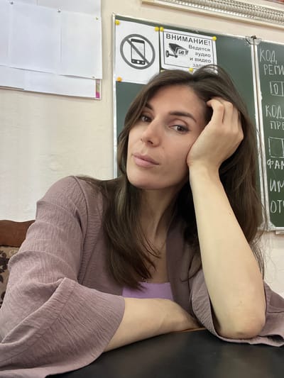 A woman is seated at a table in a classroom in Kaspiysk, Russia. She has long, dark brown hair and is wearing a light purple top and a light purple textured jacket. Her right arm is propped up, and she is resting her head on her hand, looking towards the camera. The background includes a whiteboard with writing and a sign that reads "ВНИМАНИЕ! Ведется аудио и видео записи" (ATTENTION! Audio and video recording is in progress) with an icon of a security camera. There are also papers pinned to the wall to the left of the whiteboard.