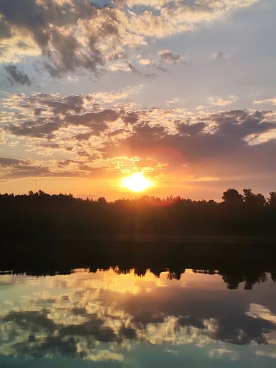 The image depicts a sunset over a body of water, likely a lake or river, in Ermolino, Russia. The sun is setting on the horizon, partially obscured by a line of trees, casting a bright orange glow across the sky. The sky is filled with clouds, ranging in color from white and grey to orange and purple, reflecting the light of the setting sun. The water reflects the sky and trees, creating a mirrored image of the scene. The tree line appears dark in silhouette against the bright sky. There are no people visible in the image.