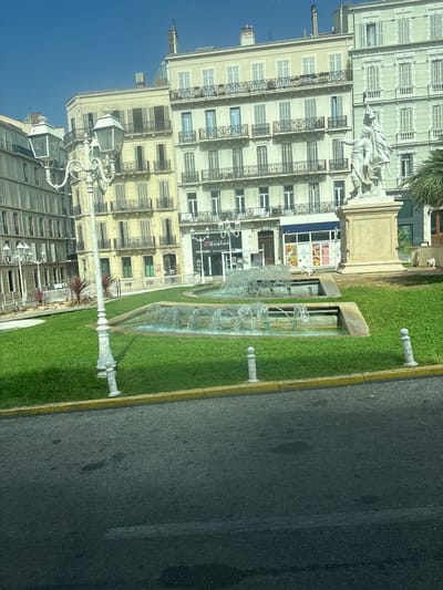 The image shows a sunny day in Toulon, France, likely in the city center given the architecture. In the foreground, a wet asphalt road curves to the right. A neatly manicured grass area with a yellow curb separates the road from a fountain. The fountain has multiple tiers, with water spraying upwards in arcs. A white, ornate lamppost stands to the left of the fountain. Behind the fountain, a statue of two figures stands on a stone pedestal. The background features several tall, multi-story buildings with classical architectural styles, characterized by arched windows, balconies with wrought-iron railings, and shutters. Several of the buildings appear to be commercial establishments, with visible signage. The sky is clear and blue, suggesting fair weather.