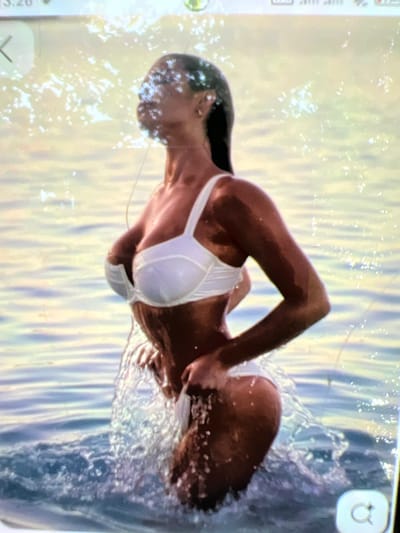 A woman in a white bikini stands in the water. Her head is turned to the right, and she is looking away from the camera. The water is rippling around her, and there are splashes of water on her skin. The background is a blur of light and color, suggesting a bright, sunny day. The image appears to be taken on a mobile phone, as evidenced by the UI elements at the top and bottom of the frame.
