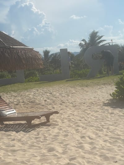 A sunny day in Bac Cam Ranh Ward, Vietnam, with a beach scene. In the foreground, a wooden lounge chair is positioned on the sand. Behind it, large concrete letters spelling out "LOVE" are visible on a grassy area. A person in a dark shirt and shorts is interacting with the letter "R," appearing to be cleaning or maintaining it. Another person, partially visible, stands near the letter "O." A thatched-roof structure, possibly part of a resort or building, is to the left, and lush green vegetation, including palm trees, forms the background. The sky is blue with scattered white clouds.