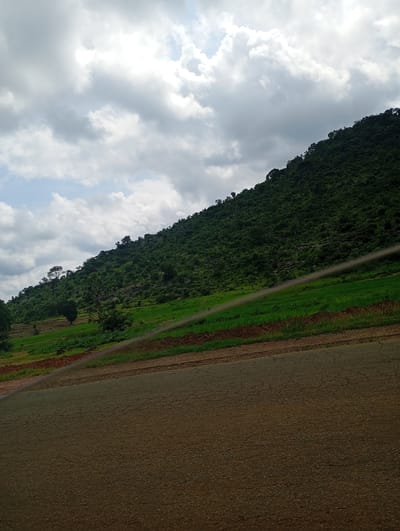 The image depicts a natural landscape with a road in the foreground and a lush green hillside in the background. The sky is partly cloudy with a mix of white and gray clouds. The hillside is densely covered with trees and vegetation, with patches of green fields and some rocky outcrops visible. The road appears to be paved and is angled across the lower part of the frame. The scene is likely captured in a rural or semi-rural setting. It is not possible to determine the specific location within Jibam, Nigeria based solely on this image. There are no people or specific actions depicted.