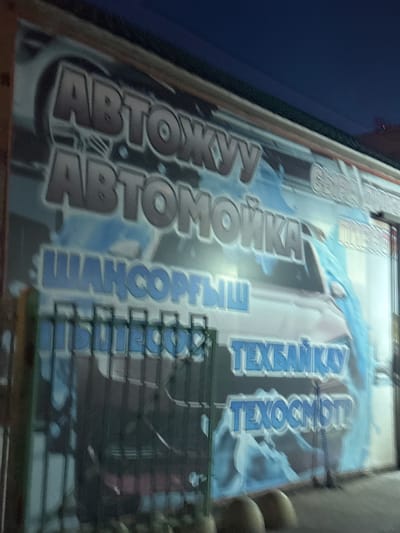 The image shows a nighttime exterior view of a car wash business. The primary subject is a large, illuminated sign with Cyrillic text and graphics. The sign features the words "АВТОКУУ" (AUTOKUU) and "АВТОМОЙКА" (AUTOMOYKA) in large, stylized, metallic-looking font. Below these, there are smaller lines of text which read "ШАНСОЛТЫШ" (SHANSOLTYSH), "ТЕХБАЙКАУ" (TEKHBAYKAW), and "ТЕХОСМОТР" (TEXOSMOTR). The background of the sign depicts a red car being sprayed with blue water. To the left of the main sign, there is a green metal fence. The overall lighting suggests it is after sunset, with artificial illumination from the business itself. The image appears to be taken from a slightly low angle, looking upwards at the signage. Based on the text, the business is likely located in a Russian-speaking region, and the context of Petropavl, Kazakhstan, aligns with this.
