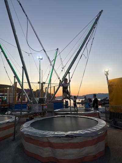 The image depicts a bungee trampoline attraction in Keramoti, Greece, during sunset. The attraction features a central metallic structure with four angled poles extending upwards, supporting bungee cords. Green LED lights are strung along the poles, illuminating the scene. A young boy is suspended in the air, attached to the bungee cords by a harness, appearing to be in mid-jump. Another child stands on a trampoline below, and in the background, several adults are visible, some observing the activity. Several circular trampolines, padded with striped orange and white fabric, are arranged on the ground. In the distance, the sea is visible, with boats docked and a faint outline of mountains against the colorful sunset sky. The overall atmosphere suggests a recreational setting at an amusement park or fair.