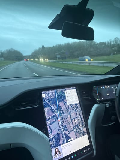 The image depicts the interior view from a Tesla vehicle traveling southbound on the A3 motorway, approaching the Liphook Road/B3004 interchange in Liphook, East Hampshire, United Kingdom. The central infotainment screen displays a satellite map showing the vehicle's position on the A3, 1.9 miles from a turn, with an estimated travel time to "02:05 (Aldershot)" and "Liphook Rd / B3004." The time displayed is 08:28. The driver's display on the right indicates a speed of 55 mph. A music player interface with the artist "Preslava" and song "Bogot Zemiom Hode" is active at the bottom of the central screen. The external environment, viewed through a slightly speckled windshield, is an overcast day with trees lining both sides of the motorway. The road surface appears damp. Oncoming traffic is visible in the distance, displaying illuminated headlights. A light-colored vehicle is also visible in motion on an elevated or parallel road segment to the right of the motorway. The vehicle's dashboard and white interior trim are visible in the foreground, along with the rearview mirror assembly mounted to the windshield.