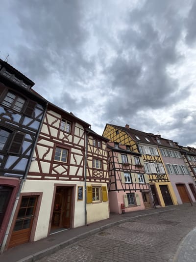 This is a low-angle, wide shot of a street in Colmar, France, featuring a row of colorful, half-timbered buildings under a cloudy sky. The architecture is characteristic of the historical district, with steep roofs and intricately patterned facades in shades of cream, red, yellow, pink, and purple. The street is paved with cobblestones, and the buildings appear to lean at various angles, creating a picturesque, slightly distorted perspective. There are no people visible in the image, and the scene is static and devoid of any overt action. The overall impression is one of charming, old-world European architecture under a somber sky, likely captured in the Petite France area of Colmar, known for its canals and traditional Alsatian houses.