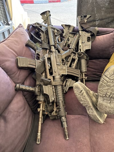 A group of five black rifles lie on a purple couch. The rifles are stacked on top of each other, and one rifle is partially resting on the floor, with the butt end of the stock resting on the couch. The lower half of a person's leg is in the photo. The person's foot is visible and is wearing a black work boot. The couch is in an outdoor setting, as the background is a paved area with a red and white striped curb. This scene appears to be in a public area in the city of Mazkeret Batya, Israel, as indicated by the latitude and longitude coordinates provided.