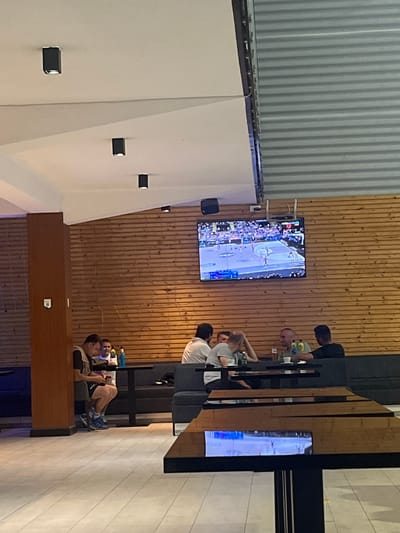 The image captures an interior scene, potentially a bar or restaurant in Pobrdje, Montenegro. The walls are a mix of corrugated metal, wood paneling, and smooth plaster. A large television screen mounted on the wood-paneled wall displays a basketball game. Several people are seated at a high table watching the game, while another television screen is on display on the other wall. Some individuals are engaged in conversations or focused on the TV, while another is using their phone. The lighting is a blend of natural and artificial. The environment is modern and functional, and the focus of the activity is the basketball game being broadcast.