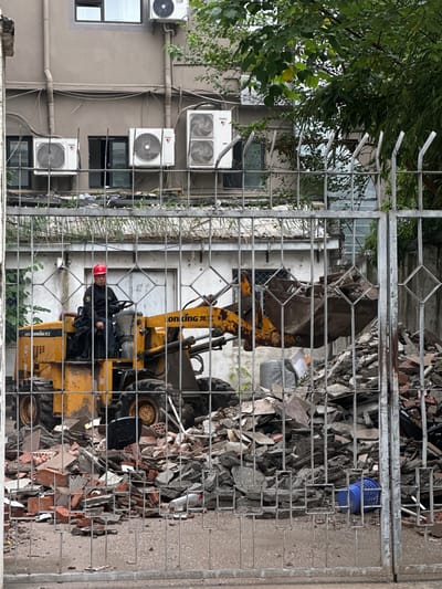 A man in a red hard hat and a black jacket operates a yellow front-end loader, which is moving a pile of debris. The debris consists of broken bricks, concrete, and other construction materials. In the background, a building with several air conditioning units is visible. The scene is partially obscured by a metal fence. The image appears to be taken in the Yangpu District of China, likely a construction or demolition site.