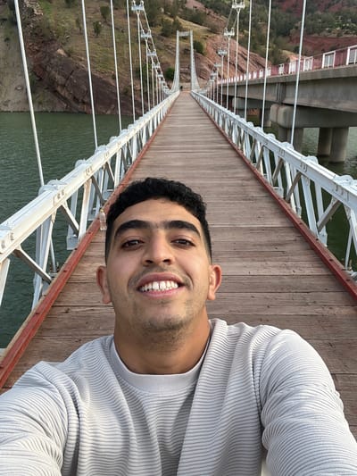 A medium-shot selfie of a smiling man with dark hair and a light gray ribbed sweater on a pedestrian suspension bridge. The bridge has a wooden deck, white metal lattice railings, and red-painted trim. Below, the bridge crosses a green-colored lake. The banks are rocky. The other side of the bridge is in the distance. The location is likely the Bin El Ouidane Dam bridge in Tabaroucht, Morocco.