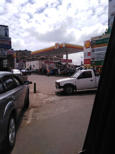 The image captures an urban street scene in Kikuyu Township ward, Kenya, centered on a Shell gas station under a partly cloudy sky. In the immediate foreground, the view is from inside a vehicle, with its dark window frame partially visible on the right. An asphalt road occupies the lower portion of the frame, leading to a paved, patterned brick entrance for the gas station.

On the left, a silver SUV is partially visible, parked or stationary parallel to the road. A black bollard stands on the paved surface between this vehicle and a white Nissan pick-up truck, which is positioned on the gas station forecourt, facing slightly right.

The Shell gas station features a prominent yellow and red canopy covering multiple fuel pumps. Several vehicles, including a dark blue car, a black SUV, a silver sedan, and a motorcycle, are situated around the pumps. Multiple individuals are visible at the pumps; one person wears an orange safety vest, suggesting a gas station attendant. Price display signs prominently feature "V-Power" fuel.

To the left of the gas station, a commercial building displays signs for "Health Services," "SCANS," "HOTEL," and "CHEMIST." Behind the gas station and the adjacent commercial structures, a multi-story building with a beige/brown facade is visible, suggesting mixed-use or residential occupation.