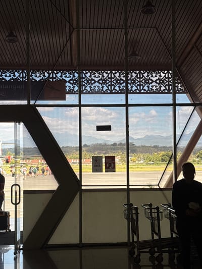 The image captures an interior view of an airport terminal in Lempeh, Indonesia, looking out through large windows towards an airfield and distant mountains. The interior features a dark, sloped wooden ceiling with exposed beams and two pendant lights. Decorative patterned screens are visible above the windows.
Through the windows, a group of people are seen walking on the tarmac, some with luggage. An airplane is partially visible on the left. The tarmac leads to a landscape of buildings and lush greenery, with a mountain range in the background under a cloudy sky. Two signs are affixed to the glass: one with a red circle and diagonal line over a person walking, and another with text. Several luggage carts are lined up on the interior side of the glass. A person stands in silhouette on the right, facing away from the camera. The floor is reflective, mirroring the interior and exterior elements.