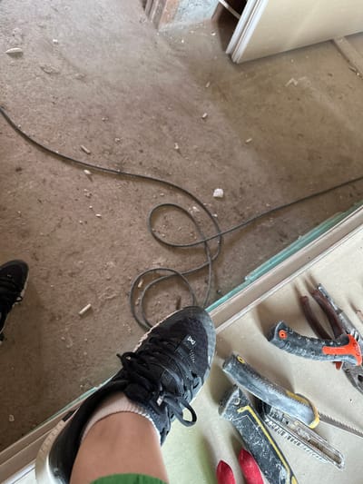 The image depicts a construction or renovation scene. The flooring is concrete covered in dust and debris. A stack of plasterboard panels is visible along one edge. Several tools, including pliers, a utility knife, and a saw, are arranged on top of the plasterboard. A black electrical cord snakes across the floor. A person wearing black athletic shoes with white soles is visible in the lower and right portions of the image. The person's foot is on the plasterboard. The specific location in Aprilovo, Bulgaria, is not discernible from the image.