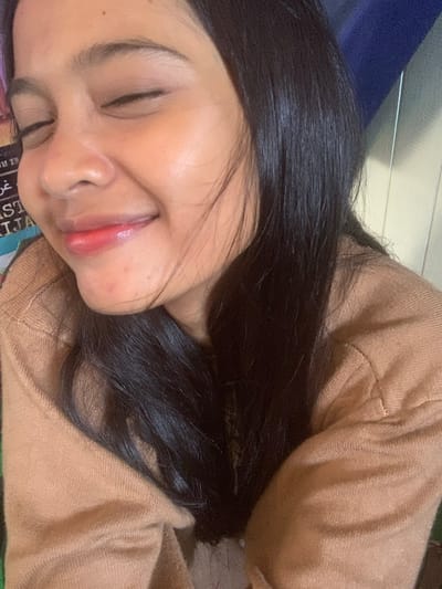 The image is a close-up, selfie-style photograph of a young woman. She has her eyes closed and is smiling. Her lips are tinted with a reddish-pink color, and she has long, dark hair that is partially obscuring her face. She is wearing a light brown, knitted sweater. The background is blurred and consists of vertical wooden planks and a dark blue object. There is also some text visible on the left side of the image, which appears to be from a poster or book. The lighting suggests an indoor setting. No specific location in Sungai Meriam, Indonesia, can be determined from the image.