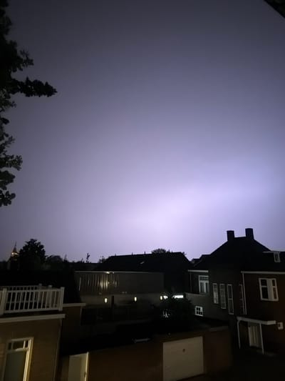 The image depicts a nocturnal scene in Boxtel, Netherlands, characterized by a bright, diffused lightning strike illuminating the sky with a purple hue. The foreground features the rooftops of several residential buildings, some with visible chimneys. A white railing adorns the balcony of a house on the left. In the middle ground, other houses and structures are visible, including what appears to be a garage door. The lighting conditions suggest it is nighttime, with the lightning providing the primary source of illumination. No people or specific actions are evident in the frame.