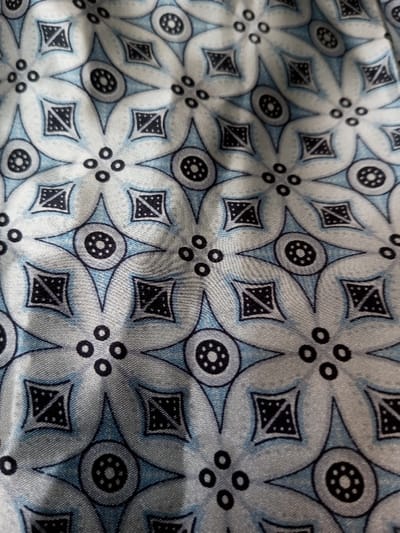 The image displays a repeating pattern with a light blue and silver-gray color scheme. The design features stylized floral shapes composed of four rounded petals, interconnected by light blue diamond shapes. Each petal contains a dark blue square with a triangular pattern inside. Where the petals meet, there is a circle filled with small black dots, or a cluster of four small dots. The fabric has a slight sheen and texture.