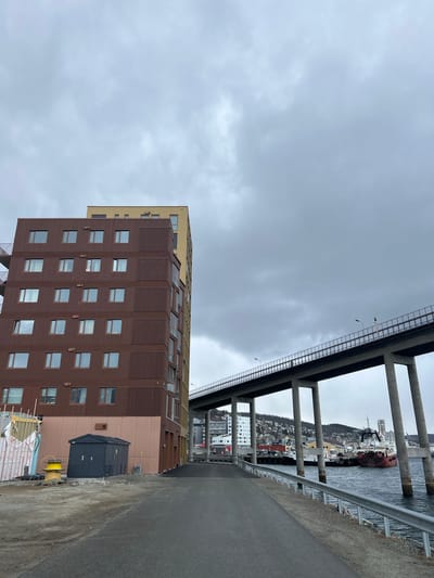 The image depicts a partly cloudy sky above a street in Tromsø, Norway. A modern brown and tan building stands to the left. The building appears to be multi-story, with numerous windows arranged in a grid-like pattern.

Above and to the right, a concrete bridge structure is visible, its support pillars stretching into the water. Beyond the bridge, the Tromsø cityscape can be seen, including buildings and waterfront activity. A road runs parallel to the water, on the right side of the frame. Construction materials are situated at the base of the building.