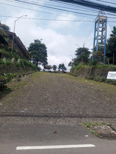 The image depicts an upward-sloping, cobblestone-paved path or road in Chori, Nigeria, bordered by vegetated embankments and structures under a partly cloudy sky. In the immediate foreground, a strip of asphalt road with a white solid line on its left edge crosses the bottom, containing a single dark red object (possibly footwear) and some debris. The paved path, dark gray and interspersed with grass and scattered litter, ascends centrally towards the horizon.
On the left, a steep, green hillside supports a long, narrow building with a brown sloped roof and an open verandah, extending upwards and to the left. A street lamp on a curved pole is positioned near this structure. On the right, another vegetated slope features a tall, blue, metal-framed water tower in the upper mid-ground. Below the tower on the right embankment, a white banner with dark, illegible text is partially visible. Further up the path, in the background, smaller trees and a small white structure with a red roof are discernible. Overhead, multiple black utility lines traverse the upper portion of the frame. No individuals or active vehicles are present in the scene.