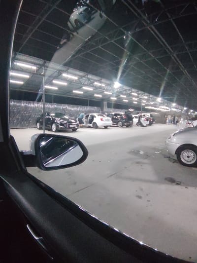 The image depicts an indoor vehicle service facility, likely a car wash or detailing center, located in Navoiy City, Uzbekistan, observed at night. The perspective is from inside a stationary vehicle, looking out through the front passenger window; the vehicle's side mirror is prominent in the lower left foreground.

The facility features a large, dark, arched metal truss roof system, illuminated by numerous rectangular fluorescent light fixtures arranged in rows. The back wall appears to be covered with a textured or reflective material. The floor is concrete, exhibiting various wet patches and puddles.

Multiple vehicles are present. In the mid-ground, from left to right: a black sedan is parked; a white sedan has its driver's side door open, with an individual leaning into the vehicle's interior; a dark SUV or crossover has its trunk open, with an individual standing near it; and another white sedan is visible further back, surrounded by additional individuals. In the far right foreground, the front passenger side wheel and fender of a silver or light-colored vehicle are partially visible. Several other individuals are scattered throughout the facility, appearing to interact with or stand near the vehicles. The scene is artificially lit, indicating nighttime operations.