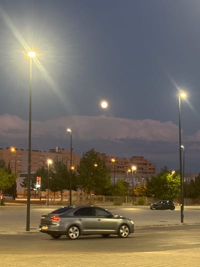 This is a full, vertical shot of a street scene at dusk in Getafe, Spain. A large, bright streetlight dominates the left side of the frame, its rays spreading outwards against the twilight sky. In the distance, a full moon is visible above a line of fluffy, pink-tinged clouds. Below the clouds, a row of buildings with many windows and lights is visible, indicative of an urban environment. Several trees with green foliage are scattered throughout the middle ground.
The foreground features a paved area, likely a parking lot or street, with two cars. A silver sedan is parked in the lower left corner, its taillights illuminated. Further into the scene, a darker colored car is driving or parked on the right side. Multiple other streetlights are present, some illuminated and some not, and a few smaller trees are also visible. The overall impression is a quiet, nighttime urban setting.