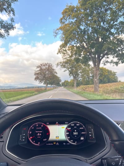 The image depicts a driver's perspective from inside a vehicle on a rural road, identified as D24B, located near Scherwiller, France. The foreground features a black steering wheel with red stitching and a digital instrument cluster. The cluster displays the time as 17:11, an ambient temperature of 17.0 °C, current fuel consumption of 8.4 l/100km, the vehicle in 7th gear (D7), a trip odometer reading 4244.5 km, and a speed of 88 km/h. An active navigation map showing the vehicle's current position and a destination marker is visible within the cluster, alongside an illuminated tire pressure warning light. The background reveals a paved two-lane road extending into the distance, lined on both sides by trees exhibiting autumn foliage. Adjacent to the road are agricultural fields. Distant hills are visible on the horizon under a partly cloudy sky with patches of blue. The headlights of other vehicles are discernible in the far distance on the road. No individuals are explicitly present.