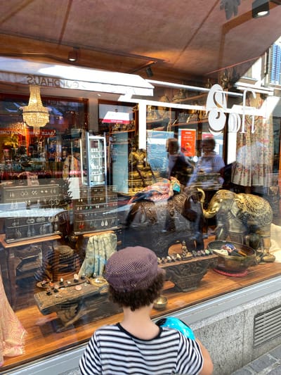 The image captures a retail storefront in Lucerne, Switzerland, displaying a range of items behind a large glass window. A young person with curly dark hair, wearing a striped shirt and a striped cap, stands facing the storefront.

Visible through the window reflection are the facade of the building across the street and pedestrian traffic. Inside the storefront, a chandelier hangs from the ceiling, and the name "GOLDAHAUS" is displayed above. A sign with the text "Sale 50%" can be seen in the middle of the window. The interior display includes a brass elephant sculpture, earrings, textiles, and other decorative objects. The display is set on a wooden surface. The overall impression is that of an upscale boutique offering a variety of jewelry and decorative items.