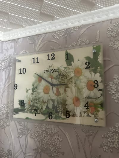 The image displays a rectangular wall clock with a floral design, featuring daisies. The clock is mounted on a wall with textured wallpaper, adorned with a repeating pattern of floral motifs and leaves. A decorative white molding runs along the top of the wall, separating it from a textured ceiling. The clock has black Arabic numerals indicating the hours, ranging from 1 to 12. The time shown on the clock is approximately 9:46. The clock itself appears to be a modern, flat-panel design. The overall impression is of an interior domestic setting, likely a living room or hallway. The image does not provide any specific information to determine its location in Yazykova Pyatina, Russia.