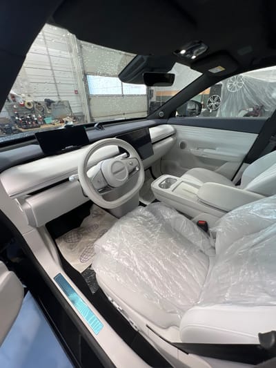 The image shows the interior of a white car, likely a new model given the protective plastic wrap on the seats and floor. The car appears to be in a garage or workshop, as evidenced by the industrial-looking doors and overhead lights in the background. The overall aesthetic of the car's interior is minimalist and modern, with a large central touchscreen and a sleek steering wheel. There are no people visible in the image. The scene is well-lit and the details of the car's interior are clear. The location is specified as Kabanikha, Russia.