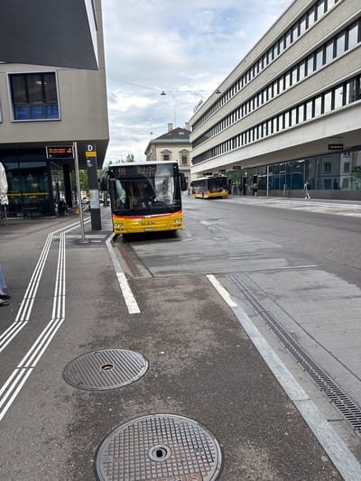 The image, rotated 90 degrees clockwise from its original orientation, depicts an urban street scene at a bus stop in Liestal, Switzerland. The perspective appears to be from a slightly elevated position looking down along the street.
In the foreground and spanning the lower left, there is a modern building with a dark, angular architectural element. Adjacent to it is a light-colored building with a large window displaying the text "DENT CARE CLINIC" and reflections of other buildings and the sky. Further along this side of the street is a bus stop sign marked with a yellow "D".
Two yellow public buses are visible, both appearing to be at or approaching the bus stop. The bus in the foreground, facing away from the viewer, is a "MAN" brand bus with the license plate "BL 208 026" and vehicle number "10418". Its destination display reads "111 Laufen, Bahnhof" with departure times "18" and "17:54". Another yellow bus is visible further down the street behind the first.
The street itself is asphalt with clear white road markings indicating bus lanes or designated waiting areas, including parallel lines and solid stop lines. A long, linear drainage grate runs along the edge of the roadway. In the lower right corner, two circular manhole covers are visible on the asphalt, one bearing a prominent "S".
To the left, a tall, multi-story building dominates the view with numerous horizontal windows creating a repetitive pattern. Its reflective surface shows reflections of the sky, clouds, and an adjacent building. Reflected signs include "SBB CFF FFS" (Swiss Federal Railways), "k kiosk", and "Mobilezone". People are sparsely visible, some walking near the buildings and others reflected in the glass facades.
The sky is overcast, suggesting daytime with diffused light. No strong shadows are present. The overall scene captures a functional public transport hub in an urban environment.