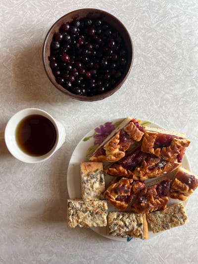 The image depicts a tabletop scene featuring food and drink. A dark-colored bowl filled with dark berries, possibly cherries or dark currants, sits to the left. A white plate holds slices of two types of cake or pie: one with a reddish jam filling and decorative crust, and the other a light-colored filling with visible seeds or nuts. A white mug filled with a dark liquid, possibly tea or coffee, is located near the bottom center of the image. The tabletop has a mottled white and grey pattern. The location is Чернышово, Russia, but no element of the picture indicates this directly. No people are visible in the image.